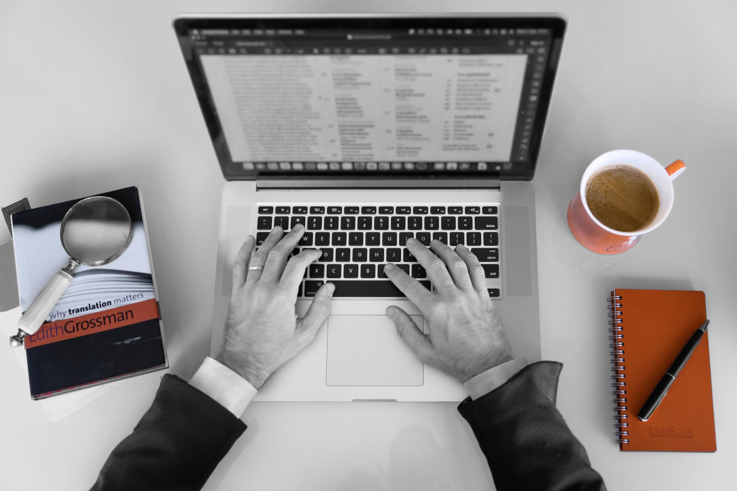 Manos trabajando encima de un ordenador, con un caf&eacute;, una libreta y un libro al lado